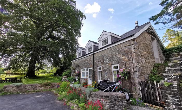 A characterful stone-built cottage with white-framed windows, surrounded by colourful flowers and climbing plants. A bike rests by a bench in the fron